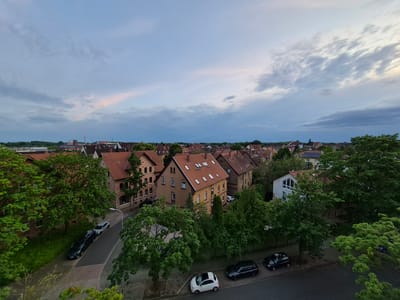 This is an aerial, wide-angle, eye-level, medium shot of a residential street in Brunswick, Germany, captured during a cloudy evening. The image focuses on a row of three-story brick townhouses with red tile roofs, interspersed with lush green trees. A paved street runs through the scene, with several parked cars, including a dark SUV, a lighter colored sedan, and a white compact car with a sunroof. The sky above is predominantly gray with streaks of white and hints of pink, indicating twilight. The overall impression is one of a peaceful, quiet urban neighborhood.