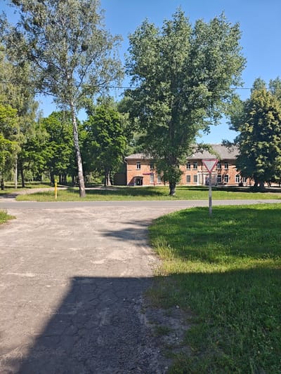 The image depicts an outdoor scene on a sunny day, likely in a rural or suburban area of Гарочыцкі сельскі Савет, Belarus. In the foreground, a cracked asphalt surface transitions to a patch of green grass. A shadow from an unseen object falls across the asphalt. Further back, a paved road curves out of view, bordered by a grassy verge. A yellow post with a green top stands by the roadside. Beyond the road, a large, multi-story brick building with numerous windows is visible, surrounded by mature trees. A triangular yield sign is positioned in front of the building, on a wooden post. The sky is clear and blue. The overall impression is one of a calm, quiet environment, possibly near an administrative building or a former communal property. No people are visible in the image.