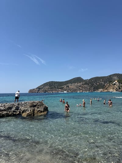 The image depicts a sunny day at a beach in Andratx, Spain. The scene is oriented vertically, showing a clear blue sky with faint wisps of clouds and a rugged, tree-covered mountain range bordering the turquoise water. Several people are visible, either swimming, wading, or relaxing in the shallow, crystal-clear water.
The foreground features a rocky outcrop, with a person standing on it, facing the water. They are wearing a white t-shirt with a number "85" printed on it and dark shorts. The water is transparent, revealing the sandy and rocky seabed below, with sunlight creating dappled patterns on the surface.
In the middle ground, various individuals are enjoying the water. Some are submerged to their waists, while others are swimming or floating. The water is calm and inviting.
The background is dominated by the dramatic mountain landscape, with some buildings nestled on its slopes, suggesting a coastal town. Several boats are anchored in the bay, adding to the sense of a popular vacation spot.
The overall mood is one of leisure and enjoyment on a warm, clear day. The bright sunlight and the serene water create a tranquil atmosphere. The orientation of the image might suggest it was taken with a phone held vertically, and the slight rotation implies it has not been corrected for its intended orientation.