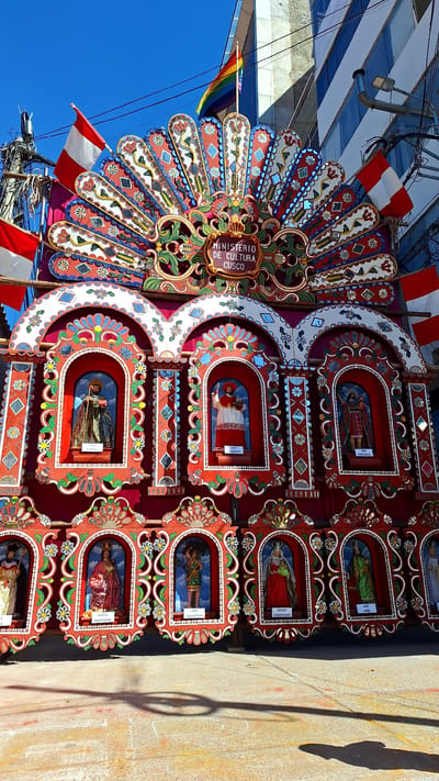 The image shows a large, ornately decorated float or altar in Wanchaq, Peru. The structure is predominantly red, with intricate colorful patterns and decorative elements. At the top, a semi-circular fan-like structure is adorned with floral motifs and small mirrored tiles. A sign in the center reads "MINISTERIO DE CULTURA CUSCO". Above this, a rainbow flag is visible flying against a clear blue sky.

The main body of the structure is divided into multiple arched niches, each containing a religious figurine. These figurines appear to be saints or religious figures, with labels below them. For instance, one niche displays "SAN ANTOLINO", another "SAN GERONIMO", and further along "VIRGEN NATIVIDAD", "SAN SEBASTIAN", "VIRGEN PURIFICADA", and "SAN JOSE". These figurines are depicted in varying attire and poses.

The float is framed by Peruvian flags on either side and is situated outdoors, likely on a street or plaza, indicated by the concrete ground surface and the shadow of a large wheel cast upon it. In the background, a modern building with large windows and some electrical wires and poles are visible, suggesting an urban setting. The overall impression is one of a cultural or religious celebration.