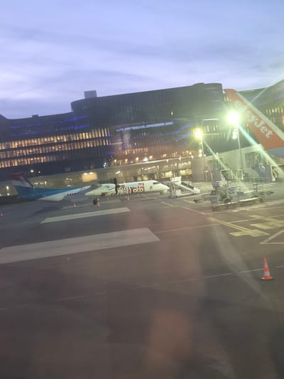 The image shows an airport scene, likely at Luxembourg Airport in Niederanven, Luxembourg. An EasyJet airplane tail is visible in the upper-left corner. Several airport vehicles and equipment are present near the aircraft. A multi-story building with numerous illuminated windows is visible behind the aircraft. An additional plane for the Luxembourg airline Luxair is visible at the bottom of the scene.