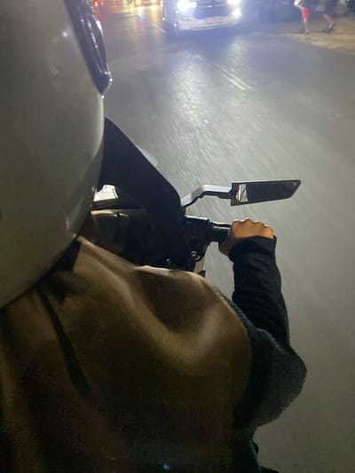 The image shows a first-person perspective from a motorcycle at night in what appears to be Macege, Indonesia. The rider's right hand, wearing a black sleeve, is on the handlebars, and a motorcycle mirror is visible. In the background, bright headlights of other vehicles illuminate a wet road, suggesting it might be raining or the road is recently wet. The overall lighting is dim, characteristic of nighttime. The scene appears to be a street or road with some traffic. No discernible text is visible in the image. The image captures a moment of travel on a motorcycle at night, with the environment lit by vehicle headlights.