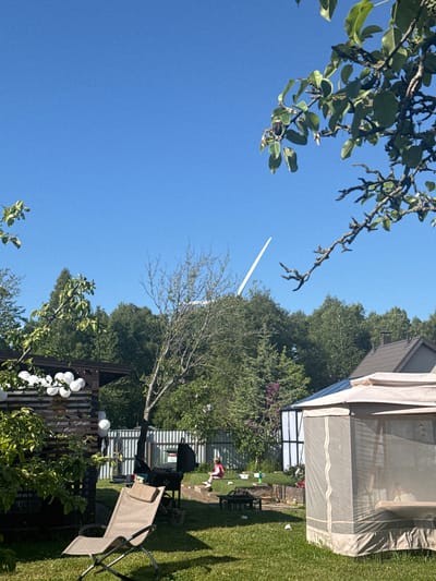 The image displays a sunny residential backyard in Šķēde, Latvia. A clear blue sky occupies the upper portion, featuring a segment of a white wind turbine blade or tower visible against it in the upper-center. Tree branches with green foliage intrude into the frame from the top-left and top-right corners.
The foreground presents a green lawn with scattered white debris, likely paper cups. An empty beige reclining garden chair is positioned on the grass in the lower-left. A black kettle-style barbecue grill is situated to the right of the chair. In the lower-right, a light-colored mesh-sided gazebo contains indistinct figures within its enclosure. A wooden pergola-like structure, decorated with white spherical lights, is partially visible on the left, obscured by foliage.
In the mid-ground, a raised wooden garden bed is located behind the barbecue grill. An individual wearing a pink top is seated on a low platform or bench to the right of the garden bed, facing away from the viewer. A white corrugated metal fence runs across the mid-ground, behind which stands a sparsely-branched tree and various other trees.
The background is characterized by a dense line of green forest trees. The brown-tiled roof and chimney of a house are visible on the far right, behind the mesh gazebo. Another building with a blue roof is partially visible behind the fence on the left side of the background.