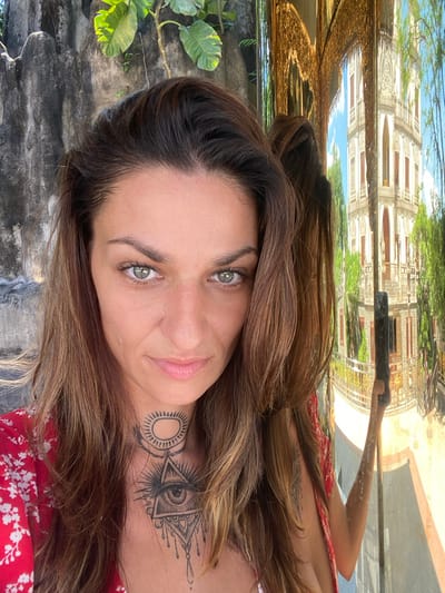 The image is a selfie of a woman, likely taken in Dĩ An, Vietnam, given the architectural reflection above her. She has light eyes and shoulder-length brown hair. A tattoo is visible on her chest and neck. She is wearing a red floral dress. The background includes weathered stonework and foliage, as well as a reflection of a building with intricate details and possibly a garden. The lighting is bright and natural. She is holding a phone to take the picture.
