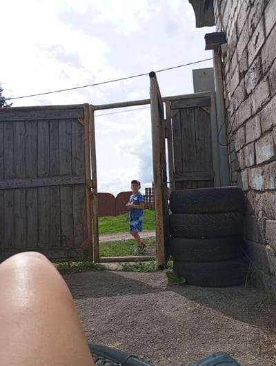 The image depicts a scene outdoors, possibly in or around the city of KODINSK, Russia, featuring a young boy standing near a rustic wooden gate. The boy, appearing to be of Caucasian descent, is wearing a blue t-shirt with a graphic design and blue patterned shorts, along with a blue baseball cap. He is looking towards the camera with a neutral expression. To the right of the boy, a brick wall runs vertically, and stacked tires are visible leaning against it. The gate is partially open, revealing a grassy area and a dirt path beyond. The foreground of the image is occupied by a person's leg, out of focus, suggesting the photo was taken from a seated or lying position. The sky is overcast with scattered clouds, and sunlight casts shadows on the ground, indicating it is daytime. The overall environment appears to be rural or semi-rural.