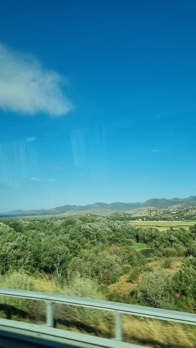 The image captures a serene rural landscape under a clear blue sky with a single white cloud on the left. The foreground features a highway guardrail with a blurred effect indicating movement, suggesting the photo was taken from a vehicle. Beyond the guardrail, a dense expanse of lush green trees and vegetation stretches into the middle ground. A small, dark body of water is visible within this green expanse, possibly a pond or a small lake. In the background, rolling hills and mountains recede into the distance, their forms softened by atmospheric perspective. The scene is likely located in the Municipal Unit of Makrichori, Greece, a region known for its picturesque natural beauty and undulating terrain. There are no people or animals visible in the image.
