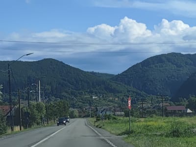 The image depicts a rural road scene in Leordina, Romania. A two-lane asphalt road curves slightly to the right as it approaches a cluster of buildings nestled at the base of densely forested, rolling hills. Two cars, a dark SUV and another dark vehicle, are traveling in the same direction as the camera. Along the left side of the road, there are utility poles with wires, streetlights, and some residential structures partially obscured by trees. On the right side of the road, a red and white stop sign is prominently displayed on a pole, accompanied by a triangular warning sign below it. Beyond the road and signs, fields of green grass and some yellow wildflowers are visible, leading up to the village buildings and the imposing green mountains in the background. The sky is a vibrant blue, punctuated by large, fluffy white clouds and some darker, more diffused cloud formations, suggesting a partly cloudy day. The overall impression is one of a serene, pastoral setting within the Romanian countryside.