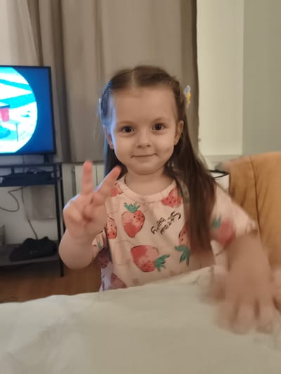 The image shows a young girl with brown hair, styled in braids with a small yellow and blue hair clip. She is wearing a light pink t-shirt with a strawberry pattern. The girl is holding up two fingers in a peace sign with her left hand. She is smiling and looking towards the camera. In the background, a television screen is visible on a stand, displaying a colorful image. There are also curtains and a chair visible. The setting appears to be indoors, likely a living room. There is no information in the image that would suggest a location in Lagny-sur-Marne, France.