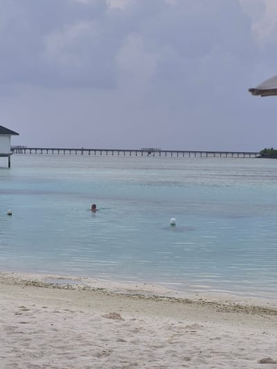 The image shows a beach scene in Huraa, Maldives. The ocean is a clear, light blue color with a small person swimming in the middle of the frame. There is a white buoy floating in the water. A long dock extends into the ocean on the left side of the frame. The beach is composed of white sand. The sky is overcast with pale gray clouds. Part of a building structure is visible at the bottom left and top left corners of the frame.