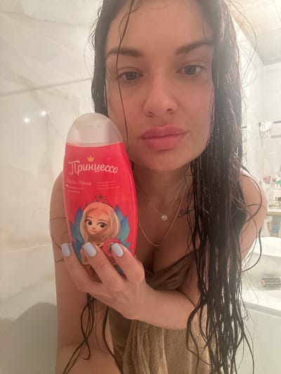 A close-up, tilted portrait shows a woman in what appears to be a bathroom or shower setting. Her dark, long, wet hair is splayed around her, indicating she has recently showered. She is wrapped in a light brown towel, visible on her shoulder and chest. Her face is turned towards the camera, with a soft expression, full lips, and dark eyes. She wears a delicate silver necklace with a square pendant and has light-colored nail polish on her fingers.
She holds a red and pink bottle of bath product in her right hand, positioned prominently in the foreground. The bottle features a cartoon illustration of a blonde princess with a crown. Visible text on the bottle is in Russian and reads: "Принцесса" (Princess), "Гель-пена" (Gel-foam), and "Облачный шоколад" (Cloudy chocolate), likely referring to its scent. Smaller text includes "Мягкая формула" (Soft formula), "без аллергенов" (hypoallergenic), and "уход и увлажнение" (care and hydration). The background consists of light-colored, possibly tiled, surfaces, characteristic of a bathroom.