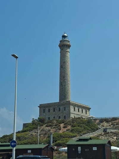 A tall, stone lighthouse stands on a hill overlooking the city of Cabo de Palos, Spain. The lighthouse has a circular tower and a lantern room at the top. In front of the lighthouse, there is a building with a green roof and a white street lamp. The sky is clear and blue, and the sun is shining. There are some people walking up the stairs to the lighthouse.
