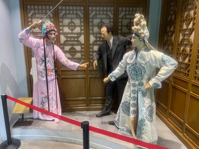The image depicts a museum exhibit, likely located in Hailing District, China, featuring wax figures representing Chinese Opera performers flanking a figure dressed in a Western-style suit.
The opera figures are in traditional costumes with elaborate headwear. One is dressed in pink with floral embroidery and the other in white and blue. The figure in the suit is dark, standing between the two opera figures and appearing to interact with them.
Behind the figures is a wooden partition with geometric patterns. A red rope barrier, supported by black posts, is set up in front of the exhibit. A sign is placed near the barrier.
The exhibit is staged on a raised platform with tiled flooring.