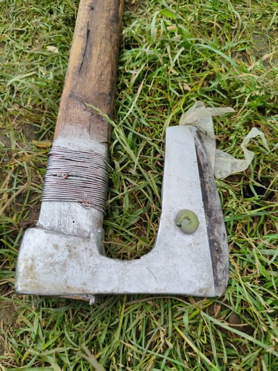 An old, weathered axe lies on a patch of green grass. The wooden handle of the axe is wrapped with a layer of thin, metallic wire for grip, showing signs of wear and tear. The metal head of the axe is a dull silver color, with a sharp blade that appears to have been used. A small, light green caterpillar is coiled on the flat surface of the axe head, near the blade. The background consists of dry, green grass, suggesting an outdoor setting in a rural or semi-rural area. There are no people visible in the image. The scene is devoid of any specific indicators that would pinpoint its location within Krushare, Bulgaria.