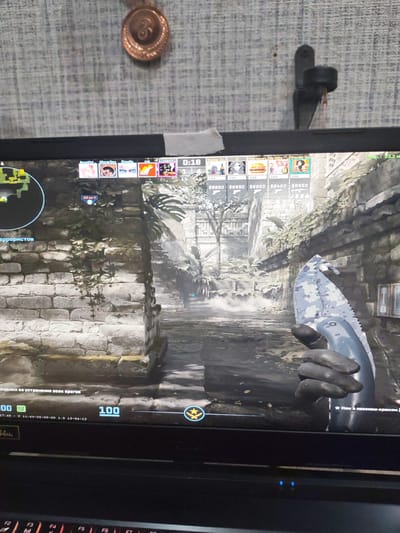 This is a photograph of a computer screen displaying a video game. The game appears to be a first-person shooter, with a player character holding a knife with a camouflage pattern. The scene depicts an outdoor environment with stone structures, foliage, and what looks like a dusty path or riverbed. In the upper portion of the screen, there are player avatars and game information, including a timer that reads "0:18". The bottom of the screen shows a keyboard, suggesting the computer is a laptop. The overall mood is intense, as expected from a gaming context. There is also a physical object, a decorative medallion, visible hanging on the wall behind the computer screen. The visible text includes game-related labels, player names, and numerical data. The date "15-04-23" is also visible, possibly indicating when the image was taken or a game date.