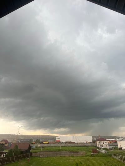 The image depicts a dramatic sky over a construction site in Solontsy, Russia. Dark, ominous storm clouds dominate the left side of the frame, creating a sense of impending weather. The right side of the image shows a more developed area with houses, some under construction, and industrial buildings. Cranes are visible, indicating ongoing building activity. The sky transitions from the dark storm clouds to a lighter, overcast sky towards the horizon, with a hint of yellow light suggesting it might be late afternoon or early evening. The overall mood is somewhat somber due to the weather, but the presence of construction implies growth and development. The weather appears to be cloudy and possibly rainy, as indicated by the dark cloud formations and the slightly muted colors of the landscape. The scene is set in a semi-urban or suburban area, given the mix of residential and industrial elements. No people are visible in the frame.