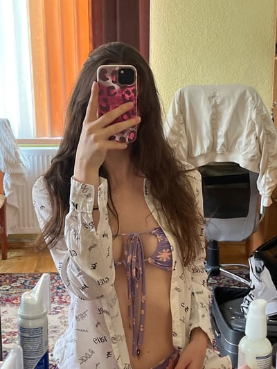 A young woman with long, brown hair takes a mirror selfie in what appears to be a bedroom. She is wearing a purple floral bikini top and an open white button-down shirt with black lettering. The room features a wooden floor, a patterned rug, a window with orange and white curtains, and a white bomber jacket draped over a chair. A bottle of nasal spray is visible on a surface in the foreground. The setting is a private indoor space, not a public location in the city of Popintsi, Bulgaria.