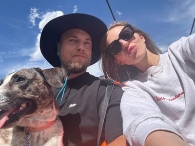 A close-up, selfie-style shot captures a man, woman, and dog on what appears to be a cable car ride with a bright blue, cloudy sky overhead. The man, on the left, wears a black hat and a black t-shirt, with a portion of a dog's head visible to his left. The dog, a brindle mix, wears a pink collar. The woman, on the right, has dark hair, wears large black sunglasses, and a light gray sweatshirt. She is leaning in towards the camera, with her lips pursed. A thin silver necklace with a script pendant is visible on her neck. The image has a slightly grainy texture, and the lighting suggests it was taken on a sunny day. The scene is outdoors and possibly at a high elevation, given the sky and the hint of a cable. The location is identified as Motički gaj, Montenegro.