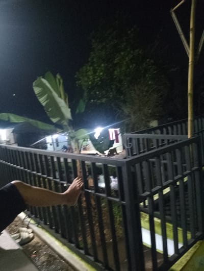 The image depicts a nighttime scene outdoors, likely in a residential area of Sukamanah, Indonesia. In the foreground, a person's left arm and right foot are visible, with the foot resting on a dark metal railing. The person appears to be wearing dark shorts or pants. Beside the person's foot on the ground are two light-colored sandals. The railing forms a barrier that separates the viewer's vantage point from the background. Beyond the railing, there are some residential buildings with illuminated windows, suggesting activity within. Large banana leaves are prominent in the mid-ground, indicating a tropical or subtropical climate. A tall, light-colored bamboo pole is visible on the right side of the frame. The overall lighting suggests it is dark, with artificial lights from the buildings providing illumination. The environment appears to be somewhat cluttered with debris or foliage on the ground near the railing.