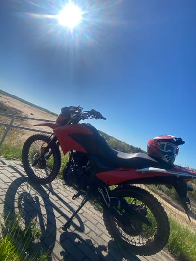The image shows a red and black dirt bike parked on a brick surface with a helmet on the back. The location is Парэцкі сельскі Савет, Belarus, as indicated by the presence of what appears to be a sand dune or quarry in the background. The sky is bright blue, and the sun is visible in the upper left corner of the frame, causing a lens flare. The motorcycle is angled in the image, and the kickstand is deployed to keep it upright. Grass grows along the edge of the brick surface. A metal fence is present in the background.