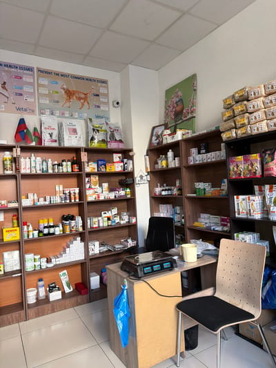 The image depicts the interior of a veterinary pharmacy located in Krumovgrad, Bulgaria. The store is well-stocked with various pet products, including food, medications, and accessories. Shelves are organized with products like Royal Canin, VetaPro, and others related to pet health. The walls are adorned with posters about pet health issues and a painting of a bird on a branch. A security camera is visible on the wall. In the foreground, a desk with a weighing scale, a yellow mug, and a chair are present, suggesting a service counter area. The floor is tiled. The overall impression is that of a functional and organized retail space dedicated to animal care.