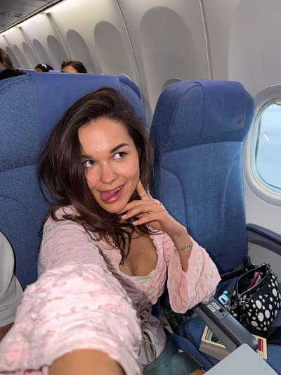 A woman with long dark brown hair is lying in an airplane seat, looking at the camera and sticking her tongue out slightly. She is wearing a light pink, textured top with a white undergarment visible. Her left hand is near her face, with a silver bracelet on her wrist. The airplane seat is upholstered in blue fabric. Above the woman, a window shows light. In the background, there is another seat with a black and white polka dot bag, a book, and a tray table. The image appears to be a selfie taken inside the aircraft. It is not possible to determine the location of the plane in Shunyi District, China, or anywhere else, based solely on this image.