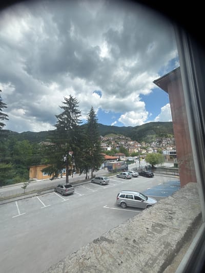 The image shows a view of a city with houses on a hillside under a cloudy sky. In the foreground, there is a parking lot with several cars, including a silver station wagon, a silver sedan, and a black sedan. There are also disabled parking spaces marked with a wheelchair symbol. In the background, there are evergreen trees and a town with buildings. The city appears to be located in a valley, with mountains in the distance. The scene suggests a residential area with a commercial presence. This is a view of the city of Devin, Bulgaria, as evidenced by the distinctive red-tiled roofs and the general architectural style visible in the distant town.