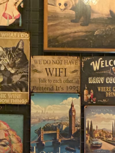 The image displays a collection of framed pictures and signs arranged on a dark, grid-like background. The top left features a partial view of a painting of a woman in a pink bikini. Below this, to the left, is a framed picture of a cat with the text "WHAT I DO" above it, and "NK WINE OW THINGS" below it. To the right of the cat picture is a sign that reads "WE DO NOT HAVE WIFI Talk to each other... Pretend It's 1995". Further to the right is a black sign with "WELCO" at the top, followed by "KANY COCO" and "WHERE G DRIN". Below these signs, a large framed picture depicts a landscape scene with Big Ben and Tower Bridge, a city skyline, and a river. To the right of this London scene is a sign featuring a Tuscan village with cypress trees and a car, with "TOSCANA" at the top and "ITALY" at the bottom. In the top center, a large framed painting shows a bear. The overall impression is a decorative wall in a restaurant or bar setting, possibly in Valletta, Malta, given the architectural style of some of the scenes.