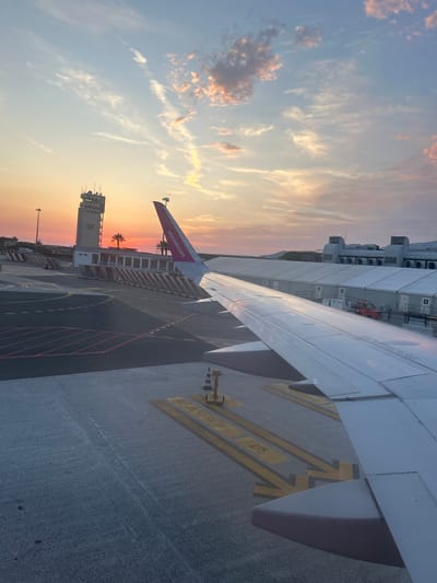 The image captures an airport scene at sunset, viewed from the window of an airplane. The sky is painted with hues of orange, pink, and blue, with scattered clouds. In the mid-ground, an airport control tower stands tall, its structure illuminated by the setting sun. Palm trees are visible behind the tower. The tarmac is marked with yellow lines, indicating taxiways or parking positions. Part of an airplane wing, white with a pink Wizz Air logo on its tail, dominates the foreground. In the background, several large, tent-like structures with ventilation units are visible. The overall environment suggests an active airport setting during the golden hour. The provided context indicates the location is Lamezia Terme, Italy.