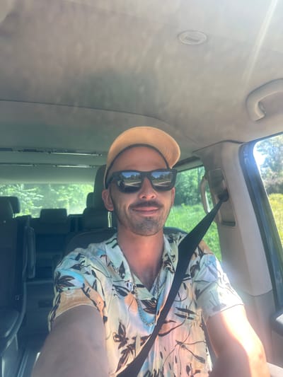 A man is sitting in the driver's seat of a vehicle, wearing a straw hat and sunglasses. He is also wearing a floral-patterned short-sleeved shirt and a seatbelt. The vehicle is likely in Bistrice, Montenegro, as suggested by the query, although this is not visually verifiable in the image. The view through the car window shows a green, possibly wooded, outdoor area. The car's interior is visible, including the ceiling, the A-pillar, and part of the dashboard.