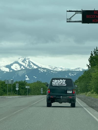 A dark green SUV with a white sticker reading "naughty by nature" on its rear window drives on a paved road. The SUV has a Russian license plate that reads "B 216 PM 41". In the background, a snow-capped mountain range is visible under a cloudy sky. A digital sign with red lettering is mounted on a metal pole above the road. The surrounding environment is lush with green trees. The scene appears to be a highway.