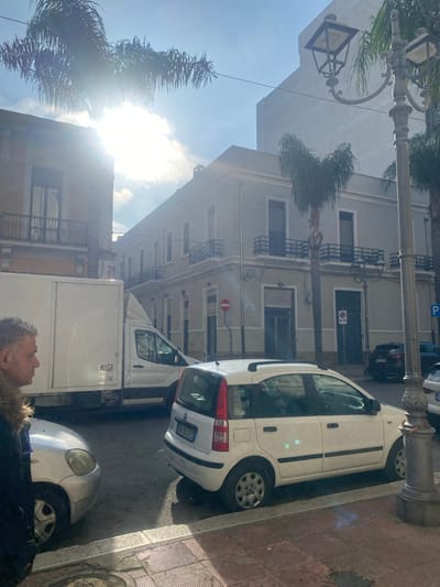 This image depicts a sunlit urban street scene in the city of Brindisi, Italy. The environment features multi-story buildings, palm trees, parked vehicles, and a pedestrian.
The sky is partly cloudy with bright sunlight, creating a significant lens flare in the upper left quadrant of the frame and casting distinct shadows on the ground.
On the left, a portion of a light-colored building with a balcony and black railing is visible. In the center-right, a prominent light-grey or beige corner building rises, featuring multiple windows, each with a small black-railed balcony on its upper floors. The ground floor displays closed shutters and entrances. A taller, lighter building is visible behind this corner structure.
Several vehicles are present. In the foreground on the right, a white Fiat Panda hatchback (Italian registration BS 151EC) is parked, facing slightly towards the camera. It is equipped with roof rails, and its front passenger-side tire appears significantly underinflated. A silver passenger car is partially visible in the lower-left foreground. A large white box truck is parked or stopped in the mid-ground on the left, partially obscuring the building behind it. Another white commercial van is visible directly behind the box truck. Further down the street on the right, a dark-colored sedan (Italian registration FZ 976CC) is parked.
Street furniture includes a decorative black lamp post with an ornate base on the right. A circular "No Stopping" sign (blue with a single red diagonal line) is affixed to a pole near the corner building, adjacent to a square "No Parking" sign. A blue rectangular parking sign with a white 'P' symbol is also visible further down the street. The sidewalk in the foreground is paved with square, patterned tiles in reddish-brown and lighter tones.
One adult male is visible in the lower-left foreground, in profile, facing right. He has short, wavy grey hair and wears a dark jacket with a prominent light-brown fur-lined collar. Palm tree fronds are visible at the top of the frame.