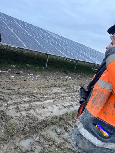 The image depicts an outdoor scene in Baldurgen, Ireland, featuring a solar panel array and muddy ground under an overcast sky. A person is partially visible on the right side of the frame, from the upper torso upwards. This individual wears a soiled orange and dark grey high-visibility work jacket with reflective strips. A dark hard hat and glasses are worn, and the right hand is raised near the face, possibly adjusting the glasses. A blue and yellow packaged item is visible in the lower right jacket pocket. The midground and background feature a large installation of multiple rows of photovoltaic solar panels, angled upwards on metal support structures, extending from the left to the right of the frame. The panels appear to have a light layer of moisture or frost. The ground throughout the foreground and beneath the panels consists of heavily saturated soil, characterized by numerous deep ruts from vehicle tires and footprints, with standing water in various depressions. Sparse green grass protrudes from the mud, and small patches of white material, consistent with frost or debris, are scattered across the ground. The sky is uniformly overcast and light grey.