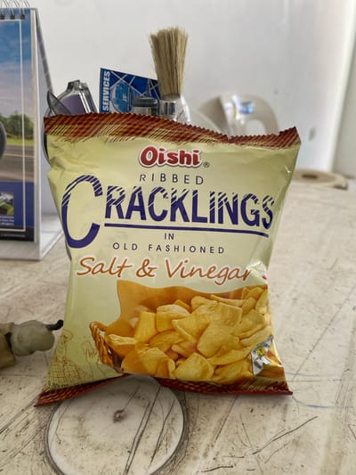 An overhead, medium shot captures a bag of Oishi Cracklings, ribbed, old-fashioned, salt & vinegar flavor. The bag is predominantly yellow with red and yellow checkered borders at the top and bottom. The brand name "Oishi" is in red lettering with a white outline, and "CRACKLINGS" is in large, dark blue, block letters with diagonal white stripes. Below this, "IN OLD FASHIONED" is in smaller, black, sans-serif font, and "Salt & Vinegar" is written in an orange script font. A stylized illustration of a basket filled with yellow, triangular potato chips is visible on the lower half of the bag.

To the left of the bag, a white calendar with a picture of a road is partially visible. Behind the bag, a small brush with a wooden handle and bristles is positioned. A blue and white card with the word "SERVICES" printed vertically is tucked behind the brush. A clear plastic object, possibly sunglasses, is also partially visible. A small, beige, human-like figurine with dark accents is placed in the lower left corner.

The items are arranged on a light-colored, textured surface that appears to be a table. The background is largely out of focus, showing a white wall and hints of other objects. The lighting suggests an indoor setting, possibly a workspace or a shelf. The scene appears to be set on a table in a store or home in San Jose del Monte, Philippines, given the context of the brand and common retail environments in the area.