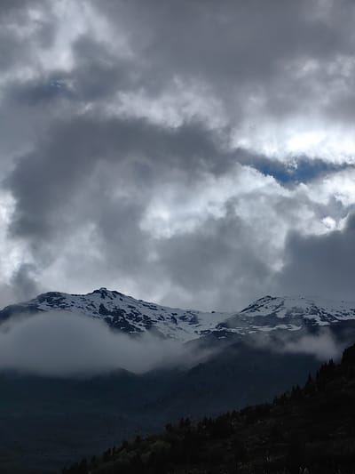 A vertical shot captures a dramatic sky over snow-capped mountains in Gemur, India. The sky is filled with heavy, dark clouds, with patches of lighter grey and white where the sun is trying to break through. Some areas of blue sky are visible through thinner clouds. Below the sky, a mountain range is visible, its peaks covered in snow. Wisps of fog or low-lying clouds obscure the lower slopes of the mountains, creating a hazy effect. The foreground shows a dark, shadowed hillside dotted with coniferous trees. The overall mood is one of impending weather change or a dramatic, moody landscape.