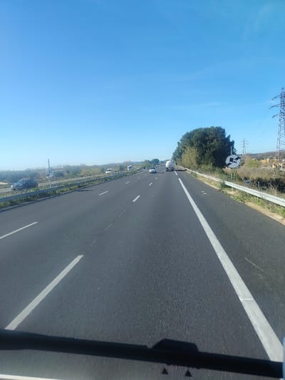 The image displays a daytime view of a multi-lane asphalt highway, likely a section of the A9 autoroute in the vicinity of Villeneuve-lès-Béziers, France. The scene is observed from the perspective of a vehicle traveling forward. The sky is clear and blue, devoid of clouds.

The highway features at least two active lanes in the direction of travel, separated by dashed white lines, and a solid white line delineating the right shoulder. Multiple vehicles are visible ahead, including a white tanker truck in the far-right lane, several passenger cars (a white sedan, a dark sedan, and other indistinct vehicles) in various lanes, all proceeding in the same direction.

To the left of the road, a metal guardrail separates the highway from a grassy embankment. Beyond this, a dark colored vehicle is stopped or parked near a white circular road sign with an indecipherable symbol. The distant left background consists of low-lying vegetation and flat terrain.

To the right of the highway, another metal guardrail runs parallel to the road, bordering a grassy verge. Immediately beyond the guardrail, a dense line of green trees screens the background. A prominent metallic lattice electricity pylon stands behind the trees, supporting power lines that extend across the landscape. Further to the right and on higher ground, several buildings or structures are visible. A black circular road sign with white symbols is situated on the right side, partially obscured by foliage.

The lower edge of the image reveals the dark outline of a vehicle's dashboard or windshield frame, casting a shadow onto the immediate foreground of the asphalt. No pedestrians or overt interactions are depicted.