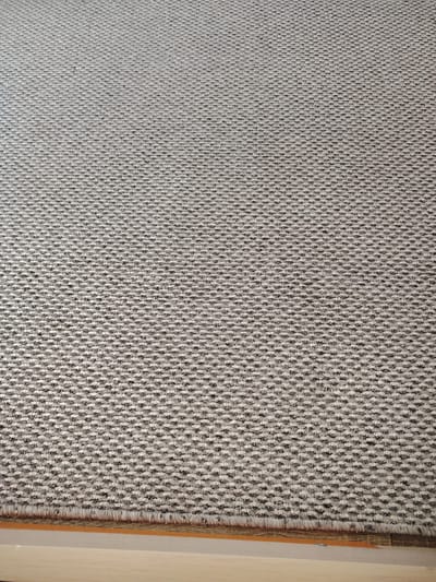 The image shows a close-up, textured view of a woven rug. The rug has a repeating geometric pattern of small, raised diamonds or clusters, alternating between light grey and dark grey threads. This creates a speckled or pixelated effect across the entire surface. The fibers appear to be short and dense, giving the rug a somewhat rough but uniform texture. The lighting suggests it is daytime, likely indoors, as there are no strong shadows indicating direct sunlight. The bottom of the image reveals the edge of the rug, where the woven pattern meets a fringe of lighter-colored, loosely hanging threads. Below the rug's edge, a sliver of a light-colored, smooth surface, possibly wood or laminate flooring, is visible. The overall impression is of a neutral-toned, modern rug, possibly a carpet sample or a detail shot of flooring. There is no text visible in the image. The location context of Putilkovo, Russia, is not directly inferable from the visual elements of the image itself.