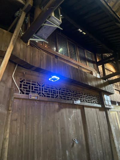 A low-angle shot captures a building's wooden exterior. A lattice-patterned window stands next to solid wood panels. A blue, cloud-shaped neon sign is positioned next to the window. A small street number sign is attached below the window. The shot includes eaves with wooden beams and rafters. A structure appears to be under construction with some building supplies.
