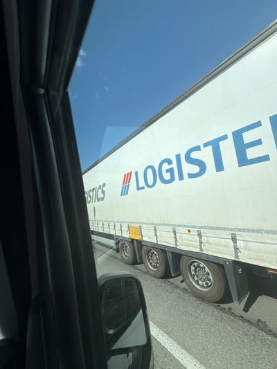 A side-view, medium shot captures a white semi-trailer truck driving on a paved road under a clear blue sky, possibly on a highway. The truck's side prominently displays the word "LOGISTICS" in large blue letters, with a red and blue logo to the left of the text. The truck appears to be in motion, with its wheels visible on the asphalt. The foreground is partially obscured by the interior of a vehicle, including a dark-colored door frame and a side mirror reflecting the surroundings. The scene is devoid of people.