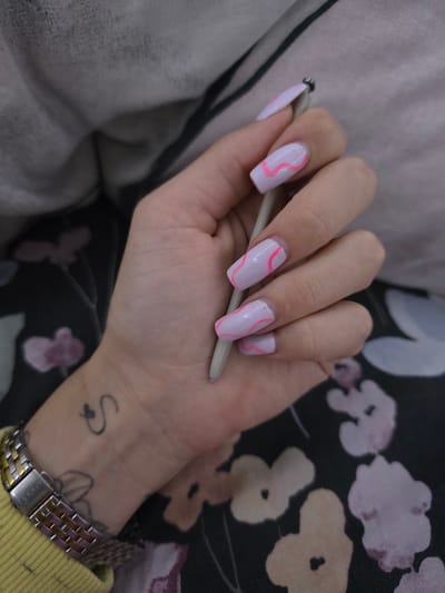 The image presents a close-up view of a person's hand holding a stylus or pen, set indoors. The hand, likely female, is positioned in the center, with fingers gently gripping a slender, light gray stylus. The nails are long and squared, painted with a light lavender or pale purple polish, and each visible nail features a distinct bright pink wavy line pattern.
On the wrist, a metallic wristwatch with a two-tone link band (silver and a rose gold or gold hue) is visible. The brand name "ESPRIT" is clearly etched on the clasp. Above the watch, on the back of the hand, there is a small black tattoo depicting a stylized cursive "S" with a small heart or loop. Part of another, larger tattoo with an abstract or floral design is partially visible beneath the watch. A yellow ribbed fabric, likely a sweater cuff or sleeve, is seen near the watch.
The background comprises soft fabrics. To the upper left, a light gray or off-white textured fabric is present. To the right and below the hand, a darker fabric, possibly black or dark green, displays an abstract floral pattern in shades of light pink, beige, and white. The setting appears casual and indoor. The provided location context for this media file is Babovo, Bulgaria.