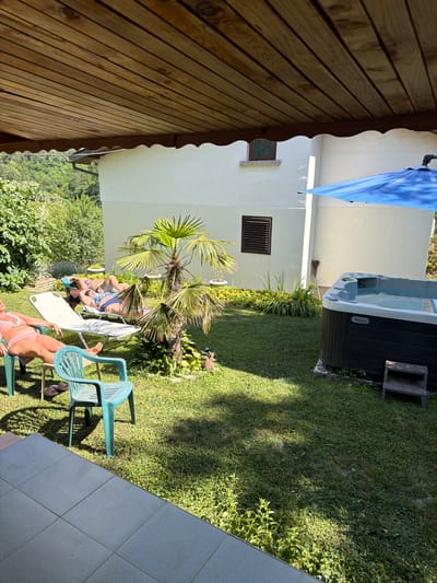 In a sun-drenched backyard in Chavei, Bulgaria, two women recline on lounge chairs under the shade of a wooden overhang. One woman, in a pink bikini, sits in a turquoise plastic chair, while the other, in a blue bikini, stretches out on a white chaise lounge. Nearby, a palm tree rustles in the breeze, and to the right, a dark-colored hot tub sits ready for use under a large blue umbrella. The scene is set against the backdrop of a white building and lush green trees.
