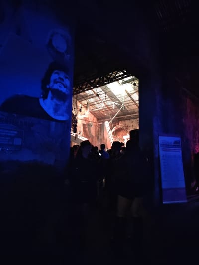 The image depicts an interior entryway leading into a larger event space within a building in Tolosa, Argentina. The foreground is dimly lit, contrasting with the brightly illuminated area beyond a central archway.
On the left, a dark wall is bathed in blue light, featuring a large, monochrome graphic of a man's head and shoulders. The man, identifiable as Diego Maradona, is depicted looking upwards with a slight smile, his features rendered in a textured, halftone-like style. Below his image, additional dark graphics or text are partially visible.
A prominent, dark archway forms the central focus, serving as a portal to the interior space. Beyond this archway, a large industrial-style building is visible, characterized by a high, corrugated metal roof supported by exposed metal trusses. Numerous artificial light sources are suspended from the ceiling and positioned around what appears to be a stage or performance area, illuminating the space with a warm, bright glow. Visible equipment includes speakers and lighting rigs, with various cables and wires running throughout.
A crowd of people is gathered within the illuminated interior, with many individuals in silhouette as they face away from the camera, suggesting movement further into the venue. Approximately 10-15 individuals are discernible near the archway, dressed in casual attire.
On the right side of the foreground, another dark wall features a vertical, illuminated sign or poster containing white text on a darker background, likely providing event-related information. The right wall also exhibits hints of magenta lighting. The floor in the foreground is dark and unadorned.