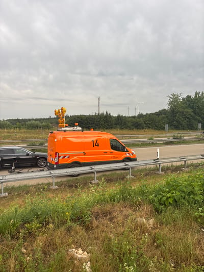 The image depicts a traffic scene on a cloudy day, likely on an Autobahn near Heidenheim an der Brenz, Germany. A bright orange maintenance van, identified by the number "14" and a logo that includes "Die Autobahn," is parked on the side of the road. The van is equipped with a tall array of warning lights and what appears to be a camera or sensor unit mounted on top. Adjacent to the van is a black car, possibly disabled, with a trailer hitch. Both vehicles are positioned behind a metal guardrail. The surrounding environment consists of a highway, grassy verges, trees, and in the distance, wind turbines and power lines are visible, indicative of an industrial or semi-rural area. The sky is overcast, suggesting a possibly damp or cool day. There are no visible people in the scene.