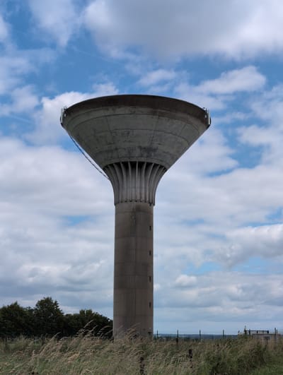 This is a full-frame, eye-level, vertical shot of a concrete water tower in Silenrieux, Belgium. The tower is the primary subject, dominating the center of the frame. It has a wide, bowl-like top that tapers down to a cylindrical shaft. The shaft is segmented and features small, rectangular windows. The base of the tower is surrounded by tall, dry grass. Behind the tower, a line of dark green trees is visible, and beyond that, a hazy landscape stretches to the horizon under a sky filled with white, cumulus clouds against a bright blue background. A metal fence runs along the lower portion of the image, separating the grassy foreground from the distant view. There are no people or animals present in the image. The lighting suggests it is daytime, likely midday or afternoon.