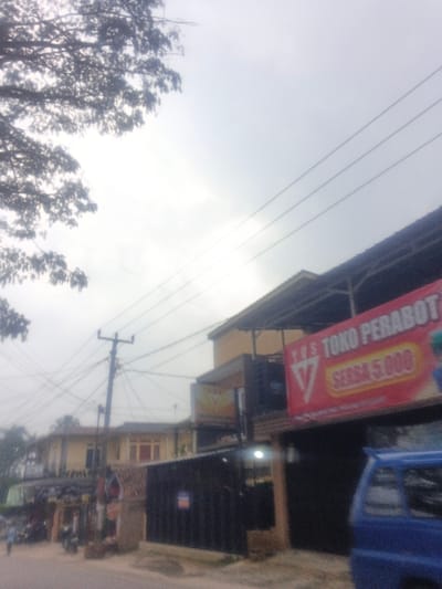 The scene depicts a street view in Dakshin Hichachhara, India, under an overcast sky. In the foreground on the right, the front portion of a blue minivan is visible. Adjacent to it, a two-story commercial building features a dark ground floor facade and a lighter upper story, possibly yellow. A large red banner is affixed to this building, displaying white text that reads "Y.O.S. TOKO PERABOT" (Y.O.S. Furniture Store) and "SERBA 5.000" (Everything 5,000), written in Indonesian.
In the mid-ground, slightly to the left of center, a utility pole stands prominently, heavily laden with a dense entanglement of black electrical wires. Multiple overhead power lines span across the upper portion of the frame. Further down the street, additional two-story buildings, primarily with lighter-colored facades and red-tiled roofs, are discernible. On the far left, a solitary individual is faintly visible walking along what appears to be a road or sidewalk. Overhanging tree branches with dark foliage occupy the upper left quadrant of the image. The ground consists of a rough road surface. The overall lighting is diffused due to the overcast conditions.