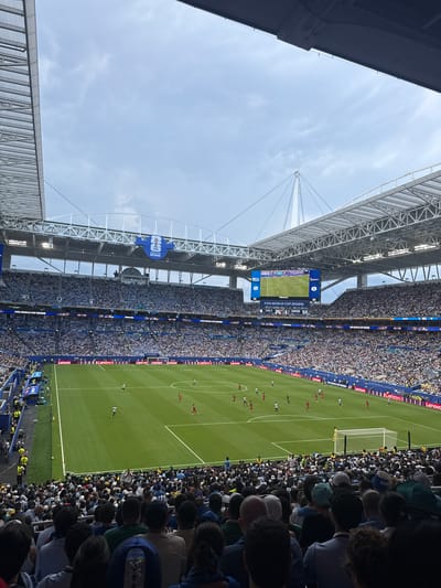 The image captures a soccer match taking place in a large, modern stadium under a cloudy sky. The stadium is filled with a dense crowd of spectators wearing predominantly white and blue attire, suggesting a supported team. The green soccer field is clearly visible, with players in red and white uniforms actively engaged in the game. Large video screens display game information, including the "FIFA WORLD CUP 2026" logo. The stadium's architecture features a prominent steel roof structure and seating tiers that extend upwards, packed with fans. The overall scene conveys the atmosphere of a major sporting event in Miami Gardens, Florida, United States of America.
