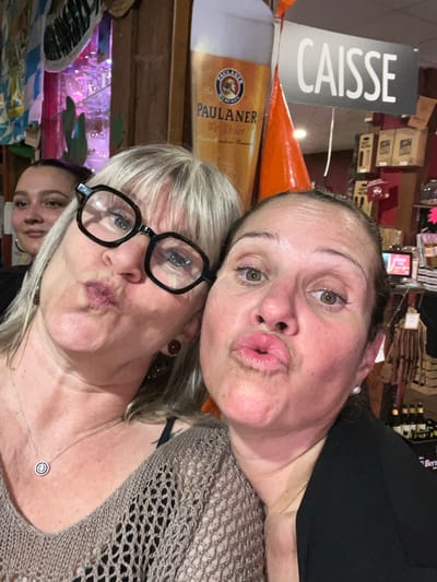 A close-up photograph captures three women in an indoor setting, identified as a retail establishment or bar in L'Étrat, France. Two women occupy the foreground, positioning themselves for the camera with puckered lips. The woman on the left has blonde hair, wears black-framed glasses, brown circular earrings, and a silver necklace with a circular pendant, dressed in a beige open-knit top. The woman on the right has darker hair, visible silver stud earrings, and wears a black top or blazer. A third woman with dark hair is partially visible in the left background, smiling subtly. The background features a large cardboard cutout of a Paulaner Weißbier glass. Above it hangs a gray sign with "CAISSE" (French for "cash register") in white capital letters, attached to an orange conical object. Shelving filled with various products, including bottles, is visible in the background, alongside decorative elements such as blue and white patterned fabric and green cactus-shaped items. The overall lighting is warm, with some purple illumination present in the far left background.