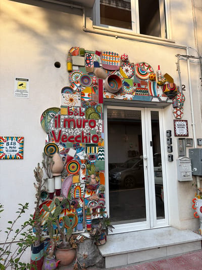 This outdoor shot shows a white building facade in Chianchitta, Italy, that features a large mosaic decoration surrounding a window. The mosaic is composed of a variety of colorful tiles and ceramic pieces, including plates, bowls, and bottles, arranged in a chaotic yet artistic manner. The words "il muro Vecchio" are incorporated into the mosaic in large red letters, with a smaller sign reading "B&B IL MURO VECCHIO" also visible below. A surveillance camera warning sign and a "NAMURI ROOMS" sign are mounted above the window, along with several electrical boxes and other wall fixtures. The window itself is white and reflects the image of a car parked inside what appears to be a garage. Plants and pots are integrated into the lower part of the mosaic, adding a natural element to the artwork. The overall impression is of a quirky and personalized building facade.