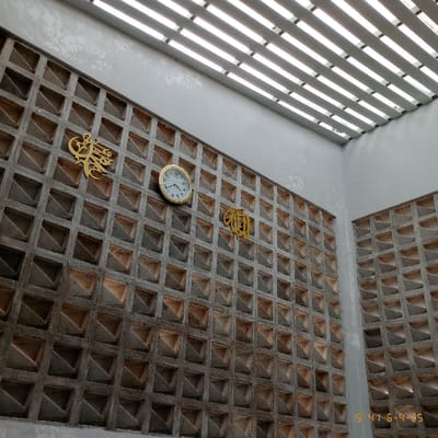 The image shows a wall made of concrete blocks with a grid-like pattern. The wall is decorated with several gold-colored ornaments and a round clock with Roman numerals. The clock is positioned on the wall, to the left of the center. The wall is seen from below, and there is a white ceiling with a grid-like structure above. The location is in Kecamatan Curug, Indonesia, with coordinates -6.266 latitude and 106.549 longitude. There are no people in the image.