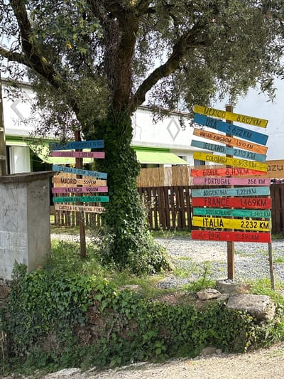 The image depicts a signpost with multiple directional signs, indicating distances to various locations from O Pino, Spain. The signpost is located amidst greenery and includes distances to local regions within Spain, like Castilla y Leon, Madrid, Valencia, Barcelona, and Castilla La Mancha. Distances to countries such as Mexico, USA, United Kingdom, Ireland, Colombia, Brazil, Portugal, Argentina, France, Germany, Italy and Korea are also indicated. The signs are colorful and of varying lengths, attached to a wooden structure, reflecting a rural or semi-rural setting. The area around the signpost includes gravel pathways, vegetation, and a glimpse of buildings in the background.