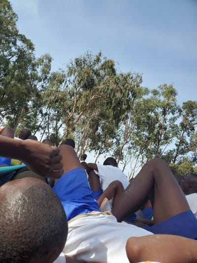 In Barakin Rafin Gora, Nigeria, a group of individuals, predominantly young men and boys, are gathered outdoors under a clear, blue sky with sparse clouds. They appear to be seated or lying down, with their legs and torsos prominently visible in the foreground. Many are wearing simple, casual attire, including white t-shirts and blue shorts. The individuals are positioned close to each other, suggesting a communal gathering, possibly for an event or relaxation. In the background, a dense canopy of trees with thin, light-colored trunks dominates the scene, with their branches reaching towards the sky, partially obscuring the view beyond. The lighting suggests it is daytime, with sunlight illuminating the area.